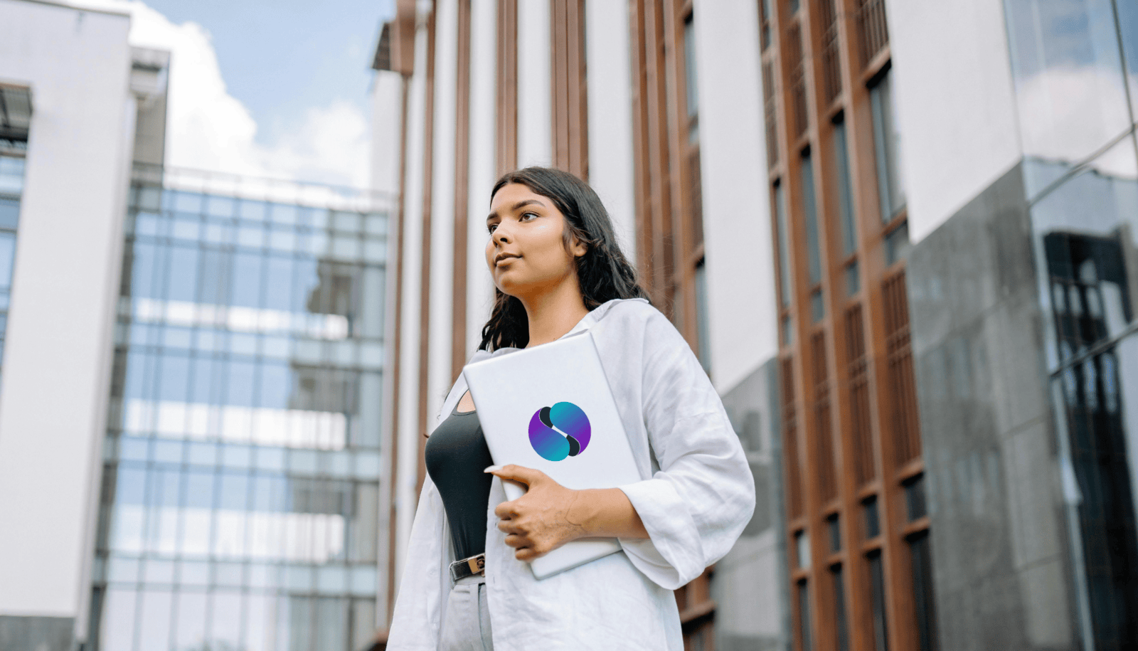 A young woman stands outside a modern university building, holding a laptop with the SPHERE logo on it. She wears a white shirt over a black top, looking ahead with determination.