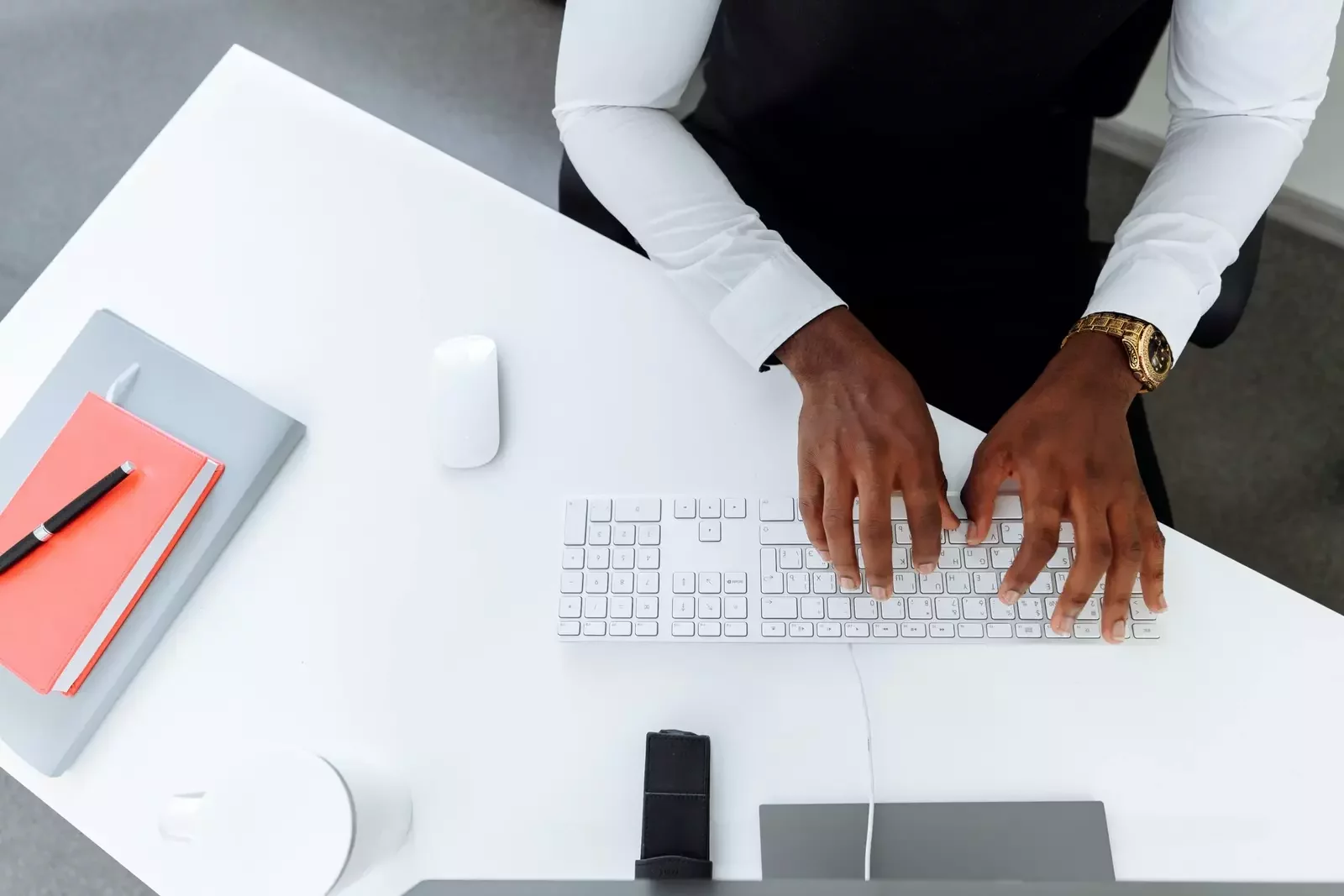 Top-down view of a person typing on a white keyboard at a minimalist white desk. The person wears a white shirt and gold watch. A notebook, mouse, coffee cup, and other office items are arranged neatly on the desk.